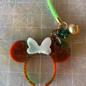 Puede incluir: Un charm hecho a mano con orejas de ratón con un lazo blanco, detalles en naranja y verde y purpurina. El charm está unido a un cordón verde con herrajes dorados, una cuenta verde y una pequeña campana dorada.