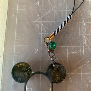 Puede incluir: Un charm de orejas de Mickey Mouse negro y verde brillante con un cordón trenzado en blanco y negro, una cuenta verde y un lazo negro. El charm está sujeto a un cierre dorado.