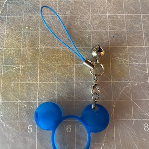 Puede incluir: Un dije azul con un diseño de orejas de ratón, una campana plateada y un cordón azul. El dije está sujeto a una cadena plateada y a un pequeño anillo plateado. El diseño de las orejas de ratón es de color azul liso.