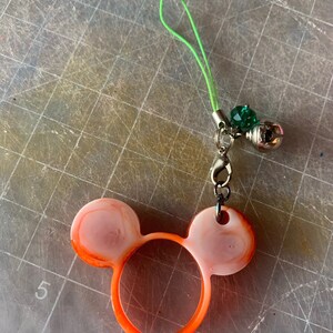Puede incluir: Un charm de orejas de ratón naranja y rosa hecho a mano con un cordón verde, una campana plateada y una cuenta verde. El charm está hecho de resina y tiene un cierre plateado.