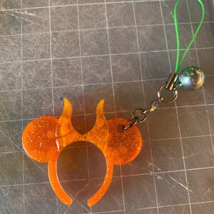 Puede incluir: Encanto de diadema de orejas de ratón de resina con purpurina naranja con un lazo verde y una campana plateada. El encanto tiene un cierre plateado y un lazo verde para la fijación. La diadema tiene un diseño en forma de llama.