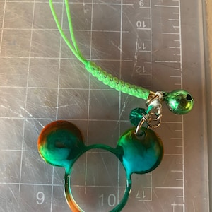 Puede incluir: Un dije de orejas de Mickey Mouse verde y naranja con un cordón trenzado verde y una campana verde. El dije está hecho de resina y tiene un acabado brillante. El dije está unido a un lazo de cordón verde.