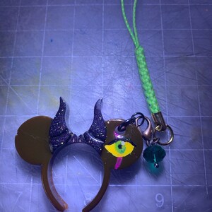 Puede incluir: Una diadema de resina marrón con cuernos de purpurina negra y orejas de ratón. Un dije a juego presenta un diseño de ojo con una cuenta verde y un cordón verde. La diadema y el dije están conectados con un anillo de metal negro.