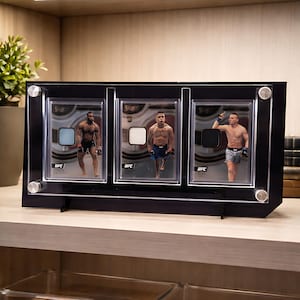 Op de afbeelding: Een zwarte acryl display case met drie UFC trading cards. Elke kaart toont een vechter en een patch. De case heeft een standaard en is bevestigd met zilverkleurige schroeven. De achtergrond is een lichtgekleurde houten plank.