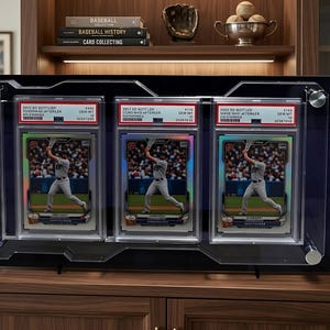 Può includere: Un espositore nero con tre carte da baseball protette. Ogni carta mostra un giocatore di baseball. Le carte hanno etichette con testo. L'espositore è su una superficie di legno. Circa 40 cm di larghezza.