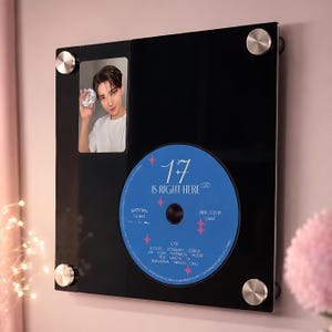 May include: A black framed display featuring a photo of a person holding a diamond-shaped object, and a blue CD with the text "17 IS RIGHT HERE". The frame has silver-coloured mounting hardware.