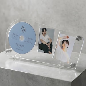Op de afbeelding: Transparante acryl displaystandaard met een cd en twee fotokaarten. De cd is lichtblauw met witte tekst en de letters "FML". De fotokaarten tonen een persoon met een wit shirt en een zwarte broek.