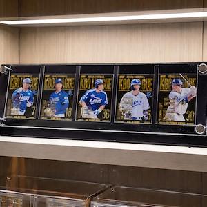 Może przedstawiać: Czarna gablota z pięcioma kartami do baseballu. Każda karta przedstawia zawodnika w niebiesko-białym uniformie. Karty są oznaczone "KBO LEAGUE" i mają złote akcenty. Gablota stoi na białej półce.