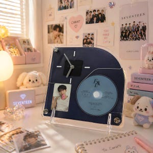 Puede incluir: Un reloj azul marino con un CD y una foto de una persona. El reloj tiene una esfera cuadrada con un recorte circular para el CD. El CD es azul claro. El reloj está sobre una superficie blanca.