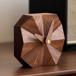 Könnte beinhalten: Eine geometrische Holzuhr mit facettiertem Design. Die Uhr hat eine dunkelbraune Oberfläche, Holzzeiger und einen kleinen goldfarbenen Punkt in der Mitte. Die Uhr ist ein dekorativer Artikel für einen Schreibtisch oder ein Regal.
