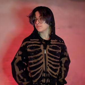May include: Black zip-up hoodie with a skeleton rib cage design in a lighter color. The hoodie has a hood and a front zipper. The person in the image is wearing glasses and has dark hair.