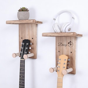 Può includere: Due supporti da parete in legno per chitarra e cuffie. Uno ha una chitarra nera, l'altro una chitarra marrone chiaro. Una piccola pianta grassa in un vaso grigio è su una mensola. Le cuffie bianche poggiano sull'altra.