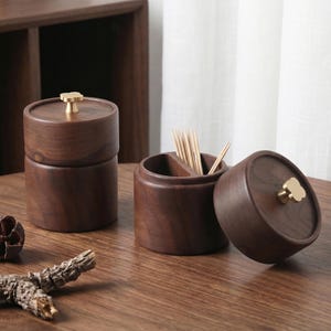 May include: Dark brown wooden toothpick holders with brass flower-shaped knobs. One holder is open, showing toothpicks. The holders are on a wooden surface. The design is simple and elegant, with a natural aesthetic.