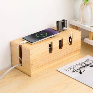 Könnte beinhalten: Ein hellfarbiger Kabelorganizer aus Holz mit drei Schlitzen, einem Smartphone, das geladen wird, und einem kleinen Ladegerät. Ein weißes Kabel ist an das Telefon angeschlossen. Ein Magazin und eine Brille liegen auf dem Tisch.