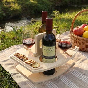 May include: A wooden wine and snack serving tray with two wine glasses and a bottle of wine. The tray holds crackers, nuts, and a bottle of wine labeled "CARO". A picnic basket with fruit is in the background.