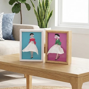 May include: Two framed art prints of a woman in a dress. One frame is white with a blue background, the other is wood with a pink background. The woman in the art is wearing a black beret, red top, and white skirt. The other woman is wearing a green top.