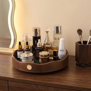 May include: A dark wooden vanity tray displays an assortment of beauty products, such as perfume bottles, skincare creams, and makeup brushes. The tray is brown with a gold emblem, and the products are arranged on a dark surface.