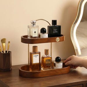 May include: A two-tiered wooden perfume organizer with gold accents. The top shelf holds three perfume bottles, and the bottom shelf holds three more. The bottles are various shapes and sizes, with clear and dark glass. A hand is reaching for a bottle.