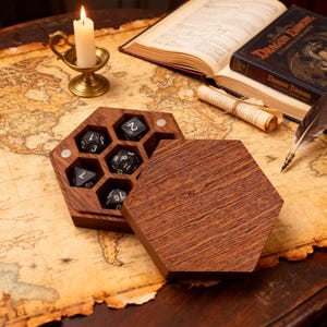 May include: A wooden hexagonal dice box with a magnetic closure, open to reveal six black dice with white numbering. The box sits on a vintage map, with a lit candle and open books in the background. The box is made of dark wood.