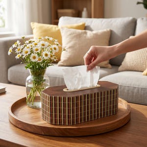 May include: A decorative wooden tissue box with a geometric design, featuring vertical stripes in shades of brown, red, and beige. The box sits on a wooden tray, with a hand reaching for a tissue. A vase of daisies is also on the tray.