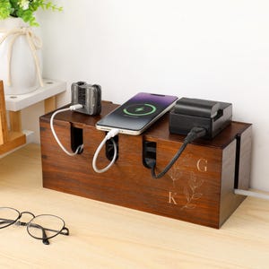 Puede incluir: Un organizador de cables de madera marrón oscuro con tres ranuras para cargar dispositivos. Un teléfono, una cámara y un cargador están enchufados. El organizador tiene las iniciales "GK" y un diseño de hojas.