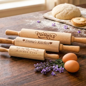 Puede incluir: Tres rodillos de madera con texto grabado, incluyendo "Happy Mother's Day", "Emma's Kitchen" y "Kitchen Love". La escena incluye huevos, lavanda y una pequeña tarta sobre una superficie de madera.