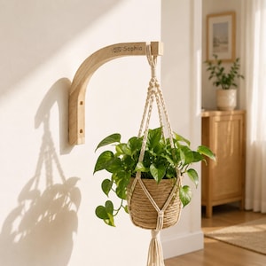 Puede incluir: Un soporte de pared para plantas de madera clara con el nombre Sophia y un pequeño icono de sobre grabados en la parte superior. Un colgador de plantas de cuerda de yute natural cuelga del soporte, conteniendo una planta potos verde.
