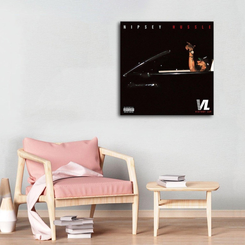 May include: A framed art print featuring the text "Nipsey Hussle" in red above a dark image of a car. The image includes a person's arm and the logo "VL Victory Lap". The print is displayed on a white wall.