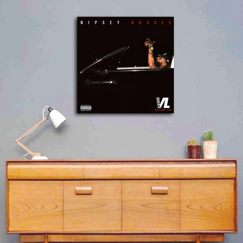 May include: A framed art print featuring the album cover for Nipsey Hussle's "Victory Lap." The artwork shows a person in a car with the text "Nipsey Hussle" in red at the top. The "VL" logo is at the bottom.