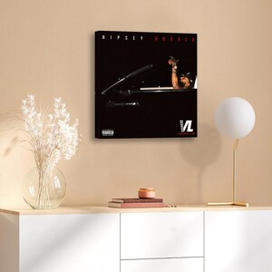 May include: A framed canvas print featuring the text "NIPSEY HUSSLE" in red at the top. The image shows a person in a car. The bottom right corner has the text "VL VICTORY LAP". The print is displayed on a beige wall.