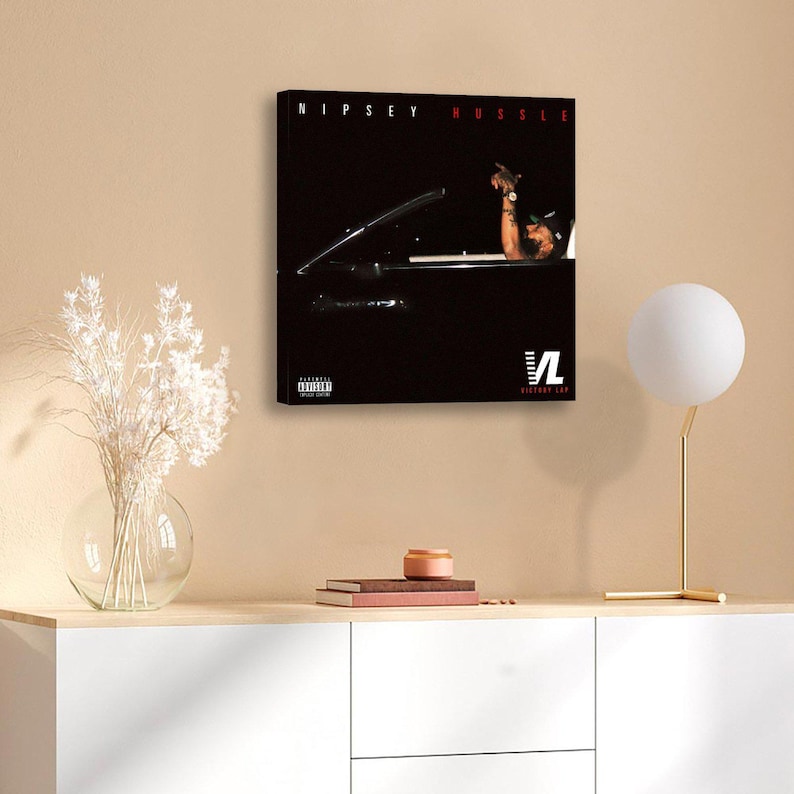 May include: A framed canvas print featuring the text "NIPSEY HUSSLE" in red at the top. The image shows a person in a car. The bottom right corner has the text "VL VICTORY LAP". The print is displayed on a beige wall.