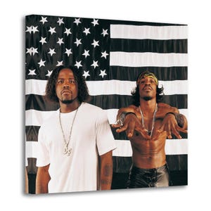 Op de afbeelding: Een canvas print met twee mannen die poseren voor een zwart-witte Amerikaanse vlag. Eén man draagt een wit t-shirt en een zilveren ketting. De andere man is ontbloot bovenlijf, met een bandana en een leren broek, met zilveren sieraden.