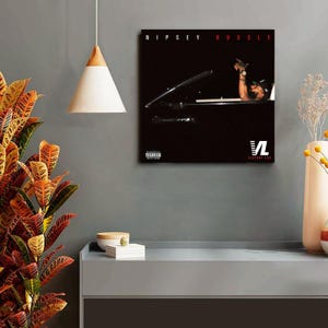 May include: A framed album cover featuring the text "Nipsey Hussle" in red against a black background. The image shows a person in a car with their arm raised. The album cover also includes the text "Victory Lap" and a parental advisory warning.
