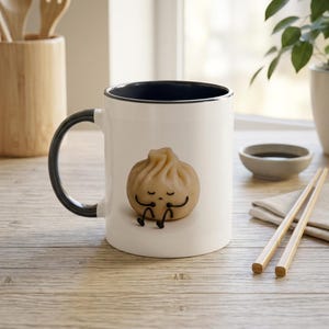 Pode incluir: Uma caneca de cerâmica branca com interior e alça pretos, apresentando um gráfico de um personagem de bolinho cozido no vapor bege com um rosto simples, braços e pernas pretos finos sentados em uma pose relaxada na parte frontal da caneca.