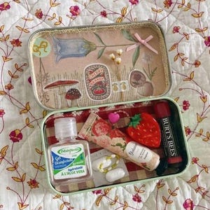 Peut inclure: Une boîte rectangulaire ouverte avec un motif floral et botanique. À l'intérieur, une petite bouteille de désinfectant pour les mains, un tube de crème parfumée à la rose, un objet en forme de fraise et un baume à lèvres Burt's Bees sont visibles. Le couvercle de la boîte présente une illustration de campanule et un autocollant "Cherry Brandy".