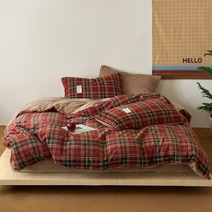 May include: A bed with a red, green, and yellow plaid comforter and matching pillowcases. The bed is made up with a tan sheet and sits on a wooden platform. A poster with the word "HELLO" is visible in the background.