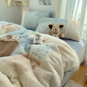 May include: A bed with a pastel-colored quilt and matching pillows. The quilt features a patchwork design with floral patterns and lace trim. A mug with the text "basics of pancake" sits on the bed, along with a stuffed dog.