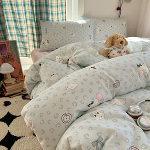 Può includere: Set di biancheria da letto azzurra con un motivo ripetuto di personaggi dei cartoni animati, nuvole e cuori. Il set include un copripiumino, federe e un cuscino a rullo. Un cane di peluche è appoggiato sopra.