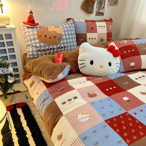 Puede incluir: Una cama con un edredón de retazos y varias almohadas. El edredón presenta un patrón de cuadrados rojos, azules y marrones, con diseños de gatos de dibujos animados. Las almohadas incluyen una almohada de cuadros azules con una cara de gato y las palabras "happy day".