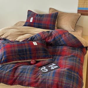Op de afbeelding: Een bed is opgemaakt met een rood, blauw en bruin geruit dekbedovertrek en bijpassende kussenslopen. Een beige kussen en een beige laken zijn ook zichtbaar. Een kleine rode appel en een tablet met de nummers 14 en 32 liggen op het bed.