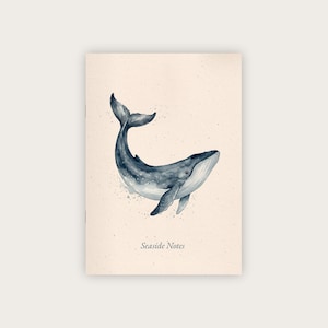 Op de afbeelding: Een notitieboek met een aquarel illustratie van een blauwe vinvis. De walvis wordt afgebeeld terwijl hij zwemt met zijn staart omhoog. Het notitieboek heeft de woorden "Seaside Notes" onderaan gedrukt. De achtergrond is een gespikkelde beige kleur.