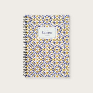 Könnte beinhalten: Ein spiralgebundenes Notizbuch mit einem weißen Einband, der mit einem blauen und gelben Muster verziert ist. Der Einband trägt den Text "meine Rezepte" in einem weißen Rechteck.