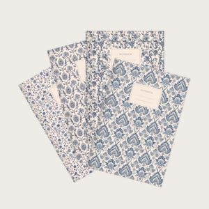Könnte beinhalten: Vier Notizbücher mit floralen Mustern in Blau- und Weißtönen. Die Notizbücher haben ein cremefarbenes Etikett mit der Aufschrift "NOTEBOOK". Die floralen Designs variieren, einige zeigen detaillierte, stilisierte Blumen, andere zartere, kleinere Blumenarrangements.