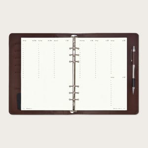 Op de afbeelding: Open bruin leren planner met een zilveren bindmechanisme. De planner is opengeklapt op een wekelijkse spread met een rasterpatroon en tijdvakken. Een zwarte en zilveren pen ligt aan de rechterkant.