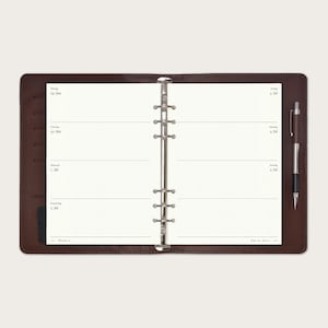 Op de afbeelding: Open bruin leren planner met een zilveren bindmechanisme. De planner is opengeklapt op een blanco wekelijkse spread met dot grid pagina's. Een pen is aan de rechterkant geplaatst. De data zijn zichtbaar op de linker- en rechterpagina's.