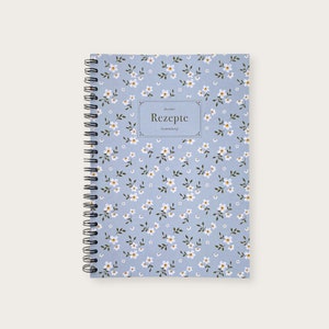 Könnte beinhalten: Ein blaues spiralgebundenes Notizbuch mit einem weißen Blumenmuster und dem Text "Rezepte" in einem schwarzen Rahmen.