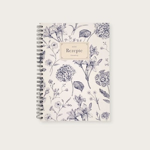 May include: A white spiral bound notebook with a floral design. The cover features a blue and white floral pattern with the text "meine Rezepte Sammlung" in a black rectangle.