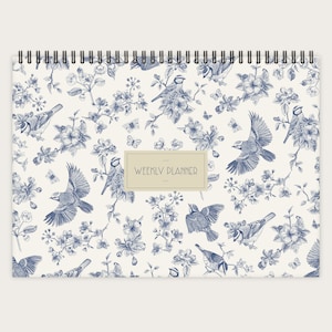 Peut inclure: Un agenda hebdomadaire blanc avec un motif floral bleu représentant des oiseaux et des papillons. Le texte "WEEKLY PLANNER" est imprimé sur la couverture.