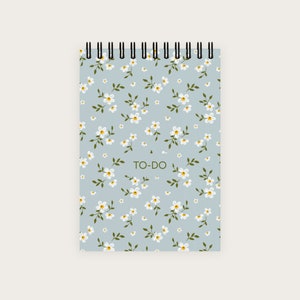 Könnte beinhalten: Ein blaues Spiralnotizbuch mit einem weißen Blumenmuster und dem Text "TO-DO" in Grün.