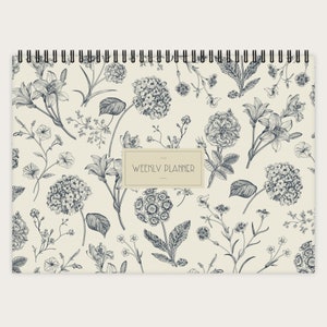 Peut inclure: Un agenda hebdomadaire avec un motif floral. La couverture présente un fond crème avec des illustrations en noir et blanc de différentes fleurs et feuilles. Le texte "WEEKLY PLANNER" est imprimé dans un rectangle brun au centre de la couverture.